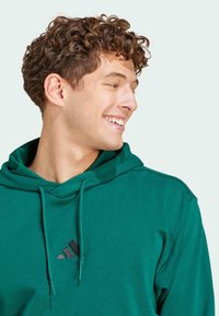 Zelená mikina s kapucňou vyrobená z mäkkého materiálu. Má kapucňu so šnúrkou a čierne logo Adidas na hrudi. Viditeľné kučeravé vlasy.