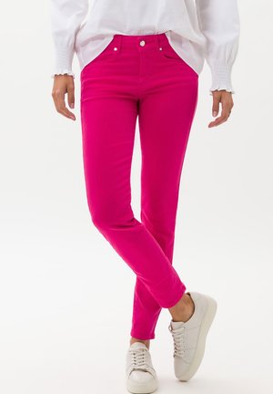Personne portant un pantalon slim rose vif, des baskets blanches et une chemise blanche à manches longues avec poignets smockés, debout jambes croisées.
