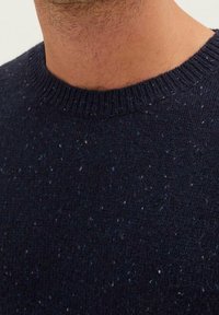 Närbild av en mans hals och bröst som bär en mörkblå fläckig stickad tröja med ribbad rund halsringning.