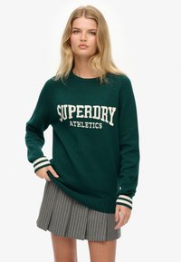 Donkergroene gebreide trui met een witte "SUPERDRY ATHLETICS"-logo, gestreepte ribgebreide manchetten en een losse pasvorm. Gecombineerd met een gestreepte rok.