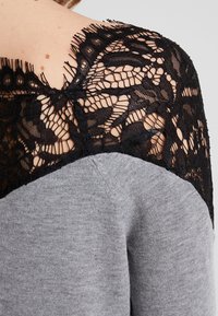 Pull en gris avec un dessus en dentelle noire sur les épaules, mettant en valeur des motifs floraux délicats et une texture douce.