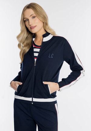 Kobieta o długich blond włosach w granatowym dresie z białymi i czerwonymi paskami, z rękami w kieszeniach, pod spodem w paskowaną koszulę.