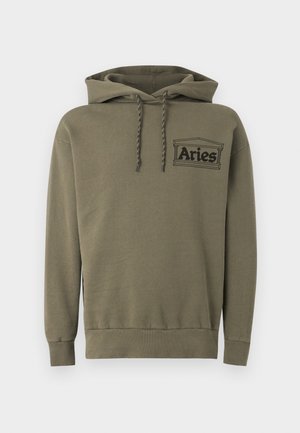 Jeune homme portant un sweat à capuche olive Aries et un pantalon patchwork assorti avec des baskets, debout les mains dans les poches devant un fond blanc.