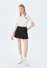 Junge Frau trägt ein weißes T-Shirt mit Katzengrafik in der Tasche, einen schwarzen Faltenrock mit Gürtel, weiße Socken und weiße Sneakers, vor einem weißen Hintergrund stehen.