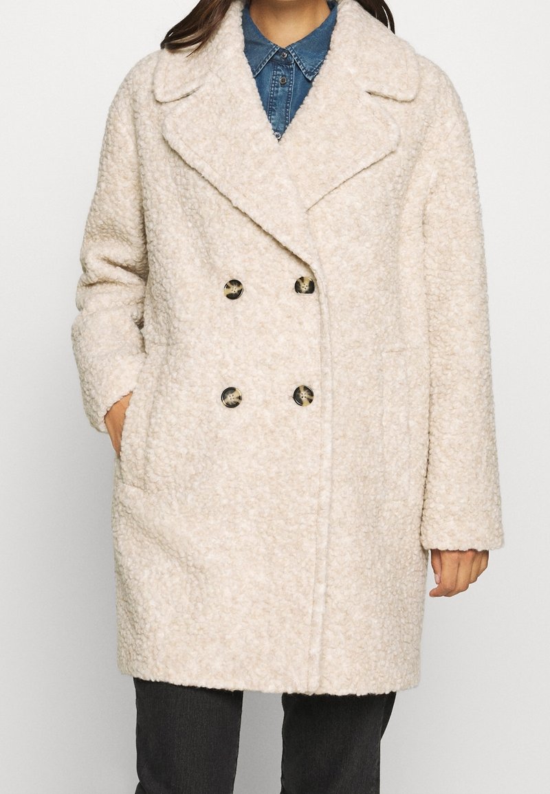 Femme portant un manteau en laine texturée crème boutonné doublement, par-dessus une chemise en denim bleu et un pantalon noir, les mains dans les poches du manteau.
