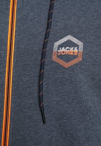 Mörkblå hoodie i texturerat tyg, med en orange och vit hexagonal logo. Svarta snoddar med orange detaljer.