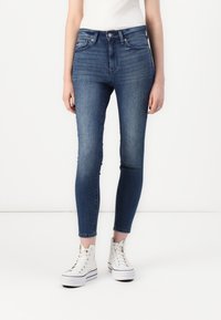 Visoke plave traperice od denima, sa slim fit krojem, minimalističkim dizajnom i krojem do gležnjeva, uparene s bijelim tenisicama.