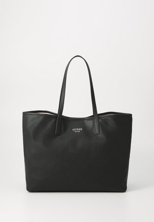 Sac cabas en cuir noir avec deux longues anses et logo « GUESS EST 1981 » centré sur le devant.