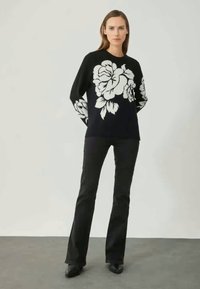 Mustapaita, jossa on suuri valkoinen kukkakuosi edessä ja hihoissa, yhdistettynä kapeisiin mustiin housuihin ja mustiin kenkiin, neutraalin taustan edessä.