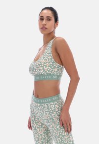 Femme portant un ensemble brassière et leggings verts et beige à imprimé léopard avec des bandes élastiques de marque Ted Baker, debout devant un fond uni.