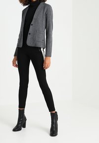 Šedé sako s jemným rybí kostí vzorem, dvěma knoflíky a klopovými kapsami, nošené přes černý rolák a černé úzké kalhoty, doplněné černými kotníkovými botami.