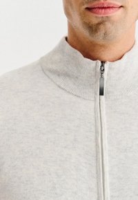 Jasnoszara rozpinana bluza z wysokim kołnierzem, z prążkowaną fakturą i gładkim wykończeniem tkaniny. Posiada metalowy zamek błyskawiczny do zapięcia.