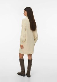 Kvinna med lång mörk hår som bär en krämfärgad stickad klänningsjumper och knähöga mörkbruna stövlar, stående och tittande åt sidan.