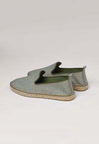 Zapatos tipo slip-on hechos de tela verde trenzada con un patrón a cuadros. Cuentan con una suela de yute y lados abiertos para poner y quitar fácilmente.