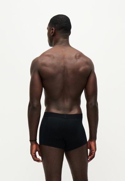 Homme musclé de dos, portant un boxer noir ajusté sur un fond clair uni.