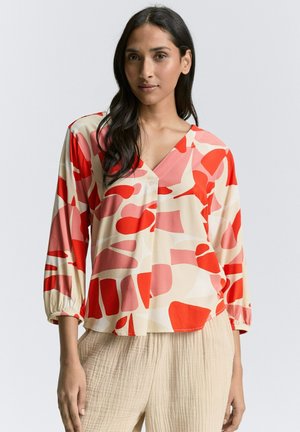 Blouse colorée avec une base crème et de grandes formes abstraites rouges et roses. Manches longues avec des poignets élastiques, décolleté en V et détail de boutons.