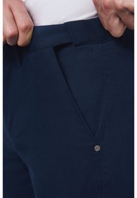 Granatowe spodnie w fakturowanym materiale, z zapięciem na guzik i subtelnym logo na pasku. Widoczne kieszenie boczne.