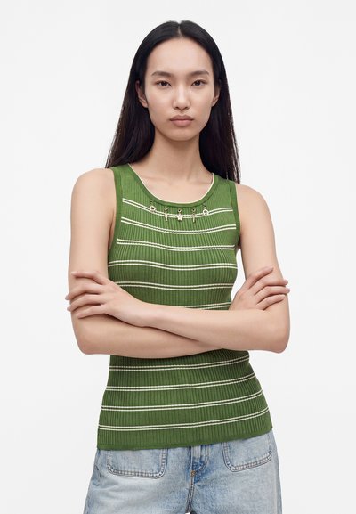 Jeune femme aux longs cheveux foncés portant un haut sans manches vert rayé et un jean bleu clair, debout, les bras croisés, sur fond blanc.