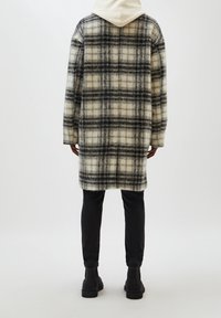 Abrigo de lana a cuadros con patrones en blanco, beige y negro, que presenta un corte relajado, hombros caídos y una capucha gris claro bajo el cuello.