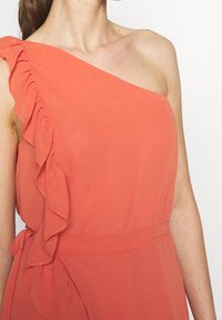 Sukienka na jedno ramię w kolorze koralowym z falbanką przy ramiączku; gładka tkanina i taliowany krój podkreślają sylwetkę.