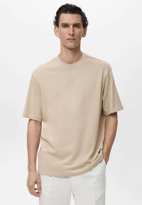 Tricou scurt din bumbac bej, cu mânecă scurtă, guler rotund, croială lejeră și detalii subtile pe laterale, asortat cu pantaloni albi.