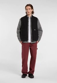 Zwarte fleecevest over een geruit overhemd en een wit T-shirt, gecombineerd met bordeauxrode cargobroek en zwarte schoenen; voorzien van ritszakken.