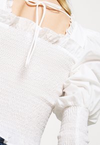 Valkoinen puffihihainen paita, jossa on rypytetty yksityskohta, röyhelökaulus ja säädettävä nauha takana. Silkkinen pinta, jossa on hieno kiilto.