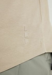Gros plan d'un tissu beige avec trois petites pièces carrées légèrement courbées disposées verticalement près de l'ourlet au-dessus d'un pantalon gris-vert.