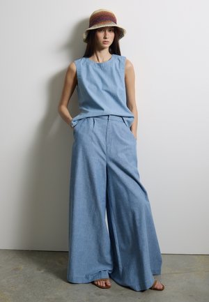 Femme portant un haut sans manches bleu clair et un pantalon large, debout avec les mains dans les poches, coiffée d'un chapeau bob crocheté multicolore.