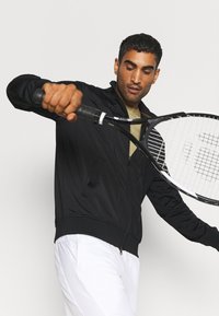 Chaqueta negra con cremallera y tejido texturizado, con costuras diagonales, llevada sobre una camiseta beige, combinada con pantalones cortos blancos y sosteniendo una raqueta de tenis.