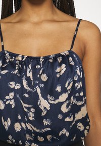 Haut sans manches bleu marine avec un col froncé, présentant un motif floral en beige et blanc. Tissu léger avec une surface légèrement texturée.