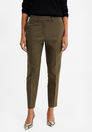 Vrouw die slank gesneden olijfgroene enkellange broek en zwarte top draagt, gecombineerd met schoenen met een slangenhuidpatroon en puntige neus, rechtstaand.