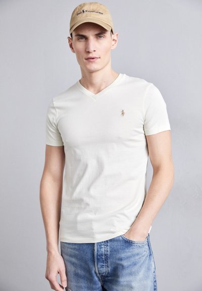 T-shirt à manches courtes crème en V, avec un petit logo brodé. Porté avec un jean en denim bleu, créant une tenue décontractée.
