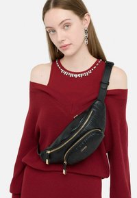 Borsa a tracolla in pelle nera con cerniere dorate e logo in rilievo, indossata sopra un maglione bordeaux a spalle scoperte in maglia.