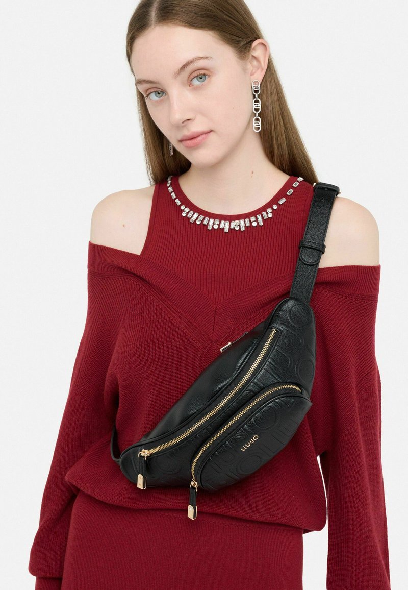 Borsa a tracolla in pelle nera con cerniere dorate e logo in rilievo, indossata sopra un maglione bordeaux a spalle scoperte in maglia.