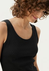 Sort tanktop med et struktureret strikmønster, bådudskæring og bølgede kanter langs ærmelukningerne og flæsen. Blødt stof.