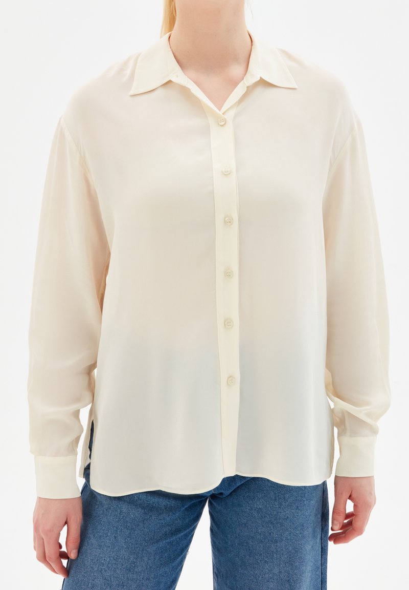 Femme portant une blouse crème à boutons à manches longues et amples, associée à un jean en denim bleu, se tenant contre un fond blanc.