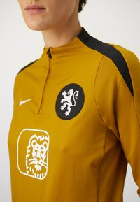 Nike sports jersey in mustard yellow with a black collar. Features a black circular patch with a white lion and a white lion graphic on the front.