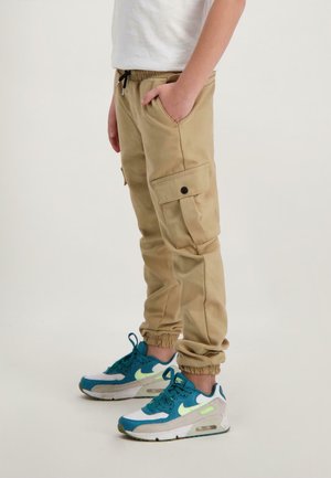 Beige cargobroek met een elastische tailleband, zijzakken en tapered cuffs, gecombineerd met blauw-witte sneakers met groene accenten.