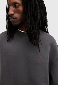 Sudadera gris oscuro con cuello redondo acanalado, que presenta un bordado de logo circular rojo en el pecho. Textura suave, corte relajado.