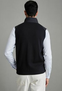 Vesta din tricot de culoare bleumarin închis, cu guler înalt, accente negre pe umeri și o textură ridată, purtată peste o cămașă cu mânecă lungă de culoare albastră deschisă.
