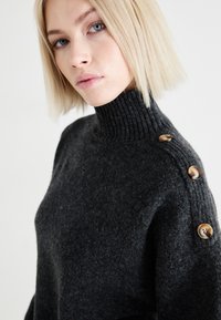 Suéter de punto de color gris oscuro con un alto cuello acanalado, que presenta grandes botones brillantes en el hombro izquierdo y una tela texturizada.