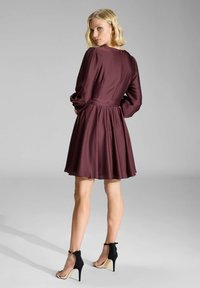 Robe bordeaux avec un corsage ajusté, de longues manches bouffantes et une jupe évasée. Portée avec des escarpins noirs à brides de cheville.