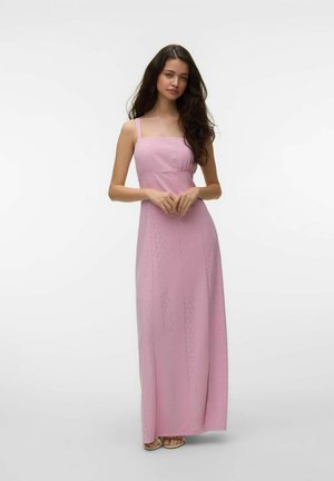 Femme aux longs cheveux foncés portant une robe longue en dentelle rose sans manches, debout les mains légèrement jointes, sur un fond clair uni.