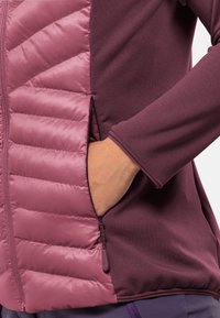 Veste violette avec une section matelassée au toucher gonflé, poche zippée sur le côté et manches lisses. Contrastes de tissus entre mat et brillant.