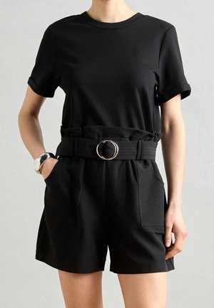 Musta lyhythihainen paita pyöreällä pääntiellä, yhdistetty korkeavyötäröisiin mustiin shortseihin, joissa on teksturoitu vyö ja suuri metallinen solki.