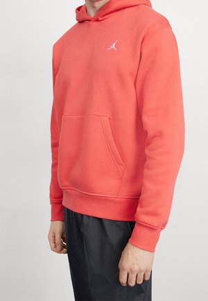 Mężczyzna w koralowo-pomarańczowej bluzie z kapturem z przednią kieszenią i małym białym logo Jumpman, w czarnych spodniach na jednolitym tle.