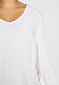 White long-sleeve blouse with a V-neckline, made of smooth fabric. Features subtle side seams and a relaxed fit.