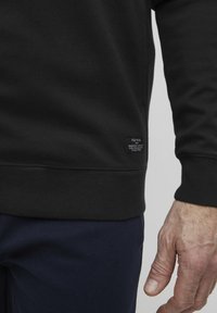 Crna dukserica s teksturiranim završetkom, s rebrastim rubom i malom tkanom oznakom sa strane. Vidljiva ruka u tamnoplave hlače.