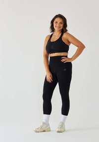 Czarny sportowy biustonosz z przednim zamkiem błyskawicznym, czarne legginsy z wysokim stanem oraz jasne buty sportowe. Gładka, elastyczna tkanina o dopasowanym kroju.